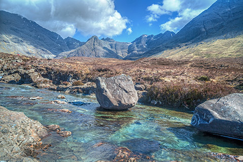 The bright blue water of the Fairy Pools flow through heather-clad moorland with the Cuillin Mountains in the background